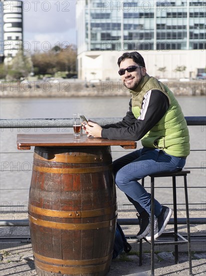 A smiling man looking at camera holding his cell phone at a bar near the river