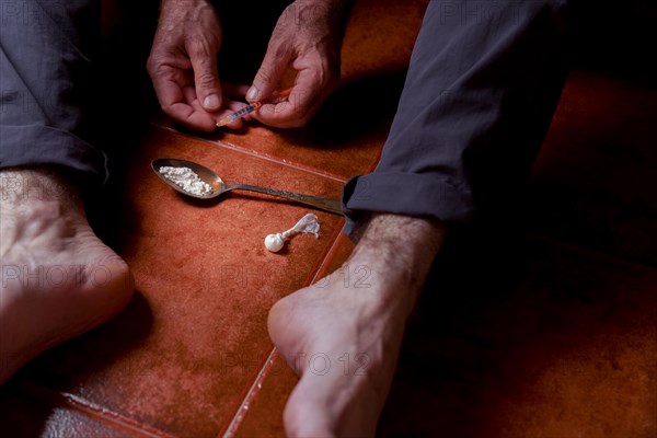 Front view of a man sitting barefoot on the floor with a heroin syringe in his hand Concept of drug addiction