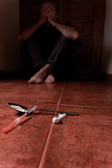 Man lying on the floor in the dark with his hands on his head desperate for drugs in the foreground Syringe with heroin and spoon