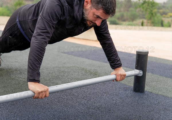 Young man dressed in black with a beard training on a barbell on the ground in an outdoor gym on a rainy day