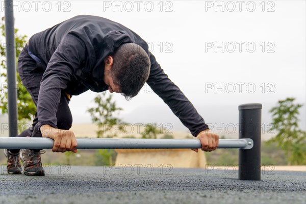 Young bearded man dressed in black training on a barbell on the ground doing stretches in an outdoor gym on a rainy day