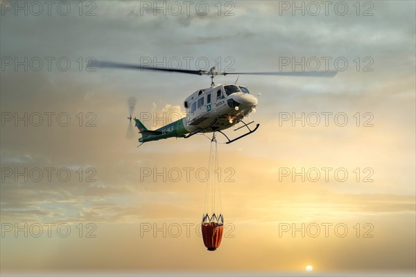 Firefighting helicopter in mid-flight dropping water with the sky red from the flames of the fire