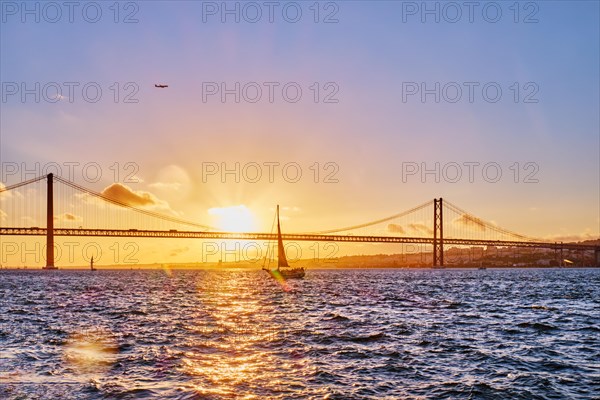 View of 25 de Abril Bridge famous tourist landmark of Lisbon connects Lisboa and Almada on Setubal Peninsula over Tagus river with tourist yacht silhouette at sunset and flying plane. Lisbon