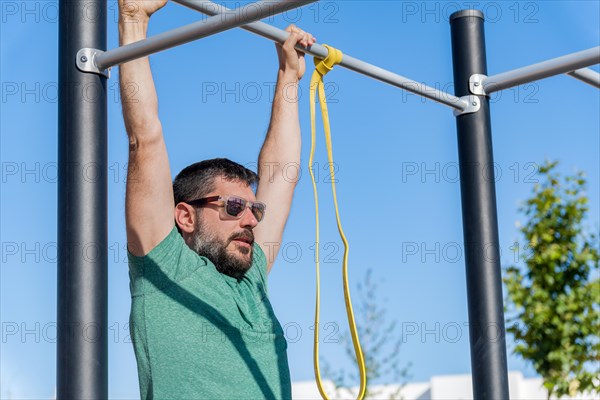 Bearded man with sunglasses seen in profile training his back by doing pull-ups on a barbell with an elastic fitness band in an outdoor gym