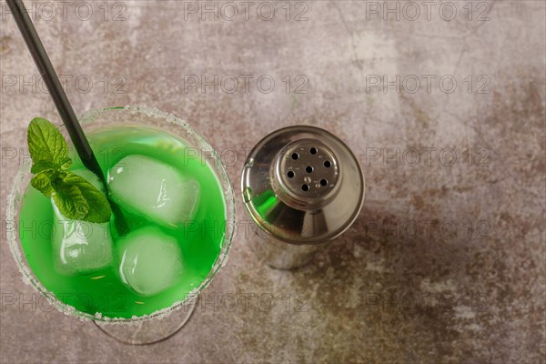 Top view of a green cocktail with mint ice and straw the edges of the glass have icing sugar and a cocktail shaker to accompany it