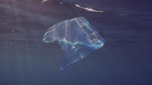 Disposable blue plastic bag floats under surface in blue water in sunrays. Plastic bag thrown into sea drifts under surface of blue water in morning sunlight