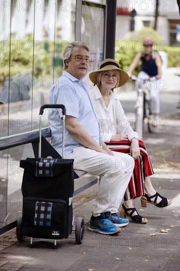 Older couple dressed in summer clothes with a trolley waiting for a bus at a bus stop