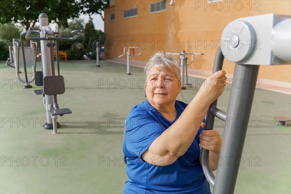 Active obese elderly woman in sportswear working out on a gym machine in a park gym while listening to music on headphones