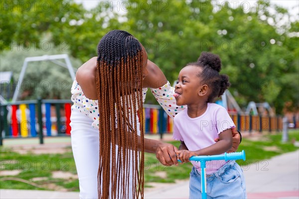 Black African ethnicity mother having fun with her daughter in playground walking with the skateboard and sticking out her tongue