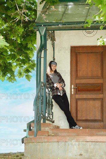 Attractive long-haired man stands on threshold of house leaning against railing in a sunny day