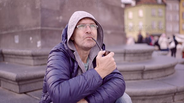 Portrait of adult man with glasses sitting in the hood on square and smoking a tobacco pipe in the Palace Square