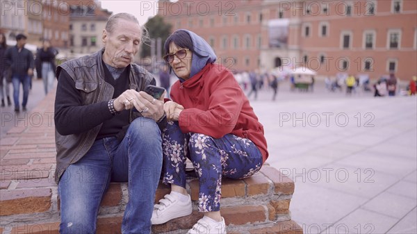 Elderly couple of tourists looking at their mobile phone are sitting in the historic center of an old European city and talking to each other. Palace Square