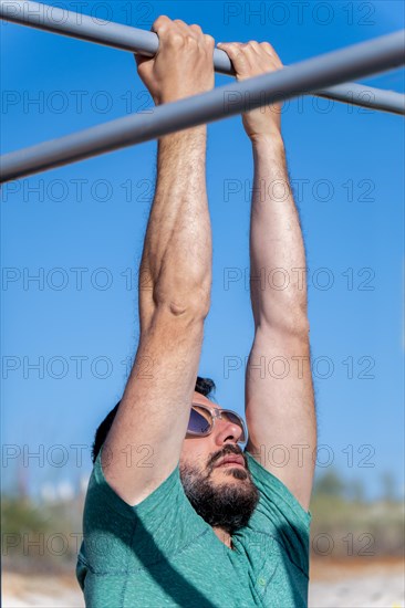 Bearded man with sunglasses seen in profile training his back by doing pull-ups on a barbell in an outdoor gym