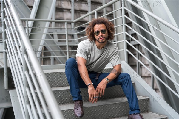 Young man with afro hair wearing sunglasses on the stairs in the city sitting