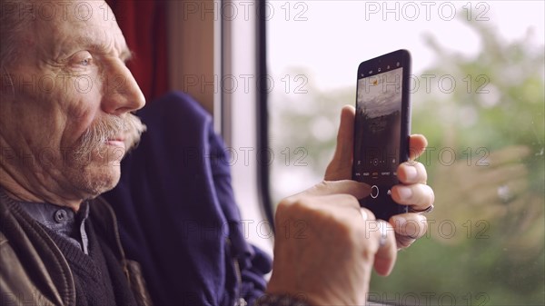 Elderly man traveling by train and taking pictures of the landscape through the window using a smartphone