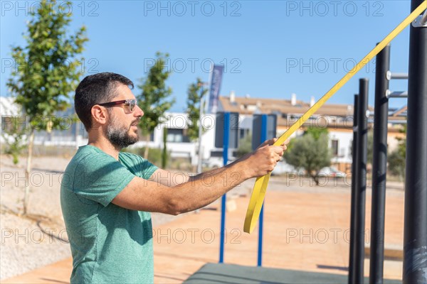 Bearded man with sunglasses seen in profile training his arms with an elastic fitness band in an outdoor gym
