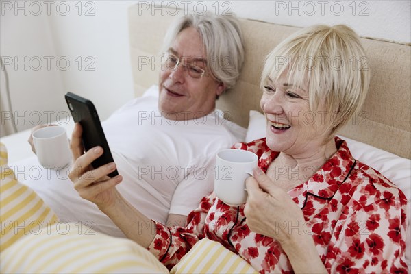 Elderly couple lying together in bed in bedroom looking at smartphone together