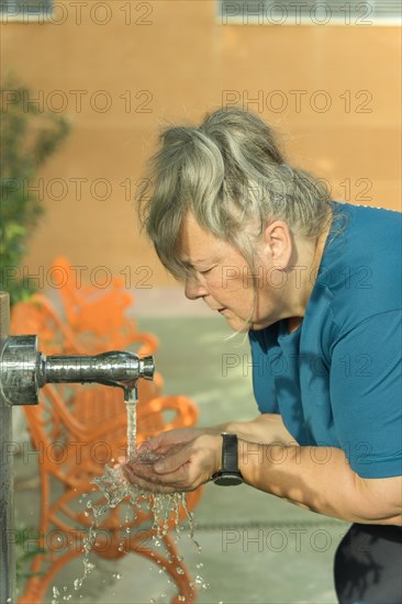 Older white-haired woman in sportswear drinking water from a fountain after workout