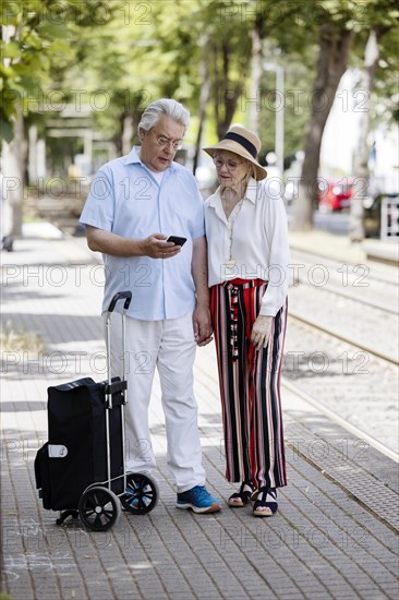 Elderly couple waiting for the tram at a stop with a smartphone in hand