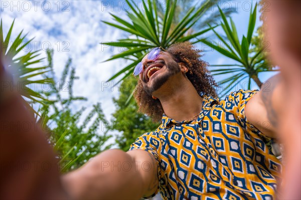 Afro-haired man on summer vacation next to some palm trees next to the beach taking a selfie with both hands smiling. Travel and tourism concept