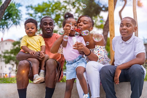 African black ethnic family with children in playground blowing soap bubbles next to trees in the park