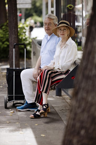 Older couple dressed in summer clothes with a trolley waiting for a bus at a bus stop