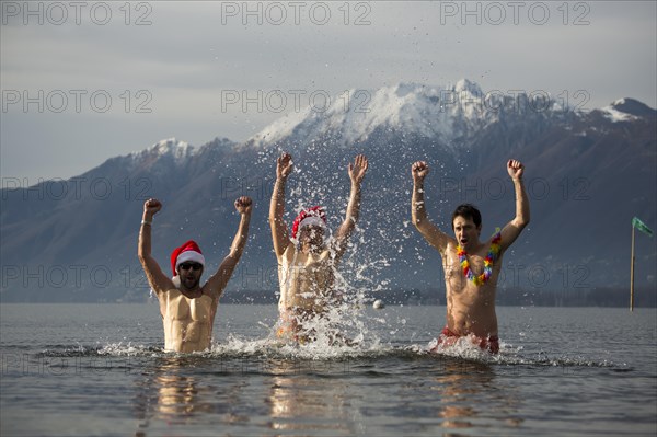 Three Happy Santa Claus with Arms Outstretched in a Cold Alpine Lake Maggiore with Snow-capped Mountain in Locarno