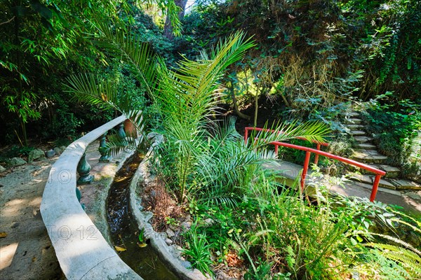 Red Chinese style bridge with wooden railings in lush greenery of asian part of tropical botanical garden in Lisbon