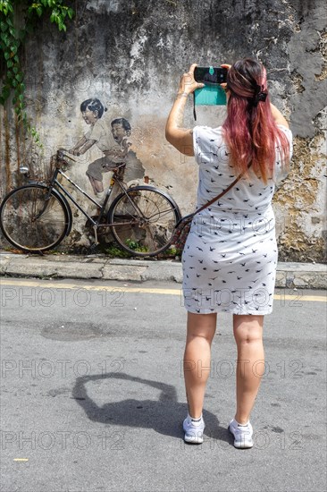 Young woman taking a photo of street art boy and girl on bicycle on a wall in George Town on Penang Island