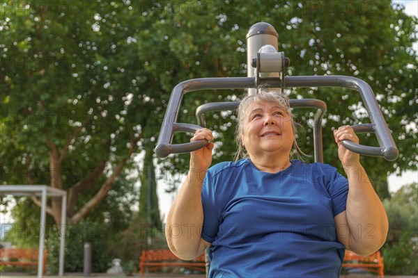 Older obese woman in sportswear exercising in an outdoor gym in a park surrounded by trees