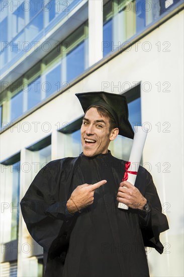 Happy graduated man wearing a bachelor gown and a black mortarboard and pointing at his diploma while looking at camera