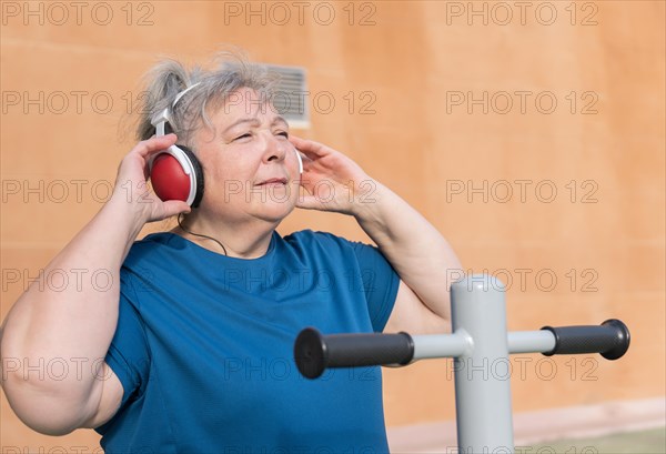 Active obese elderly woman in sportswear working out on a park machine while listening to music on her headphones