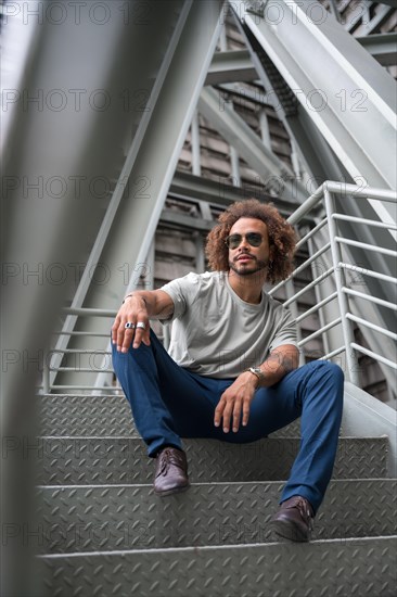 Young man with afro hair wearing sunglasses on the stairs in the city sitting looking to the left