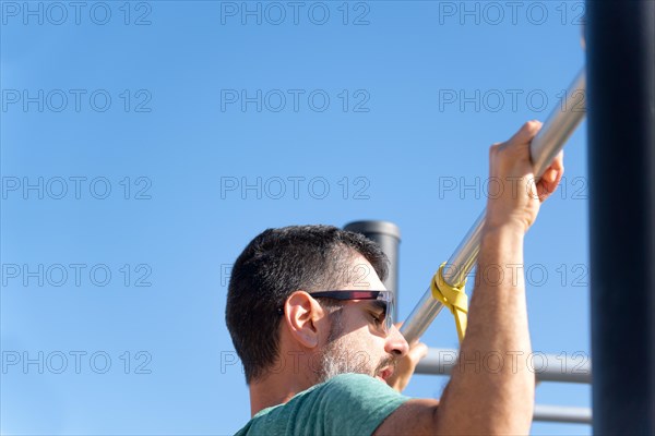 Bearded man with sunglasses seen from behind training his back by doing pull-ups on a barbell with an elastic fitness band in an outdoor gym