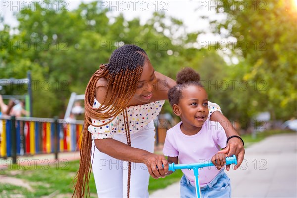 Black African ethnicity mother having fun with her daughter in playground learning to ride a skateboard in the sunset