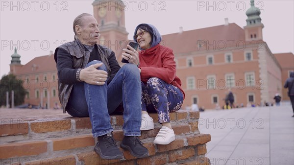 Elderly couple of tourists looking at their mobile phone are sitting in the historic center of an old European city and talking to each other. Palace Square