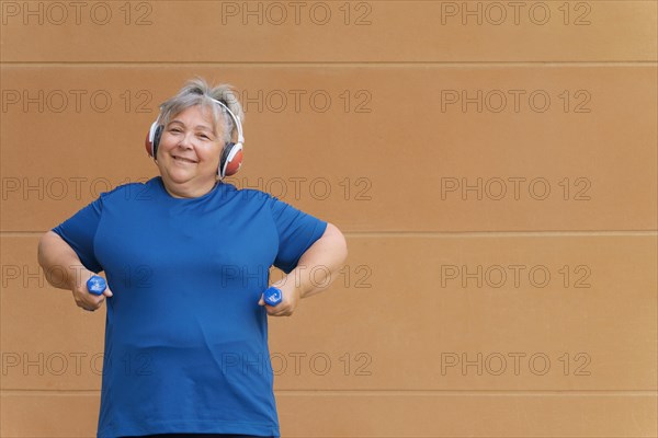 White haired obese older woman exercising with dumbbells and headphones listening to music for weight loss .isolated on orange background and copy space