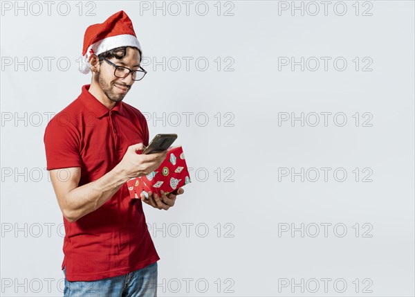 Guy in christmas hat holding gift box and smiling at smartphone. Christmas man holding gift box and telephone isolated. Handsome man in christmas hat holding gift and looking at phone
