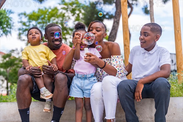 African black ethnicity family with children in playground having fun blowing soap bubbles next to trees in the park