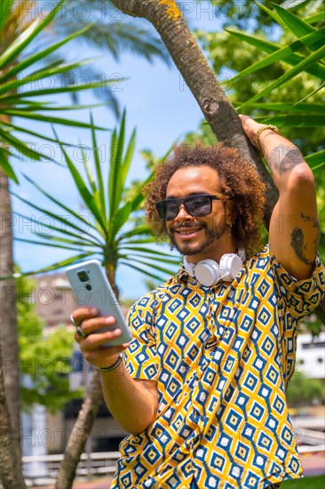 Portrait of afro-haired man on summer vacation next to some palm trees by the beach looking at the mobile. Travel and tourism concept