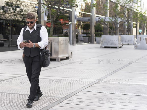 Portrait of a businessman walking on the street while he sends messages on his mobile phone