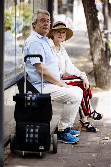 Older couple dressed in summer clothes with a trolley waiting for a bus at a bus stop