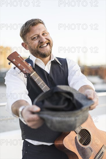 A smiling street musician looking at camera and carrying his guitar while asking for tips