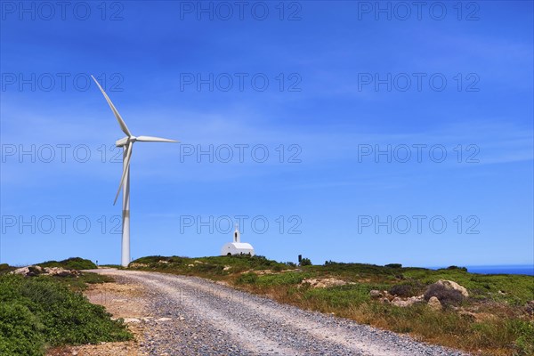 Single windmill turbine and small traditional Greek Orthodox chapel or church and road to them in colorful landscape on hilltop against blue sky with some cloulds on clear sunny summer day. Crete