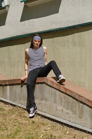 Creative long-haired man in tank top and jeans sits on railing