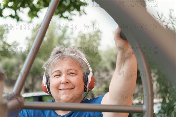 Active obese elderly woman in sportswear working out on a park exercise machine while listening to music on her headphones