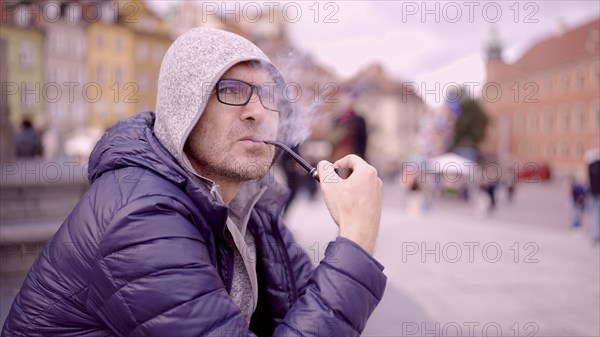 Adult man with glasses sitting on square and smoking a tobacco pipe releasing smoke in the Palace Square