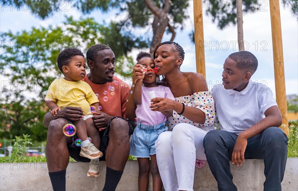 African black ethnicity family with children in playground having fun blowing soap bubbles next to trees in the park