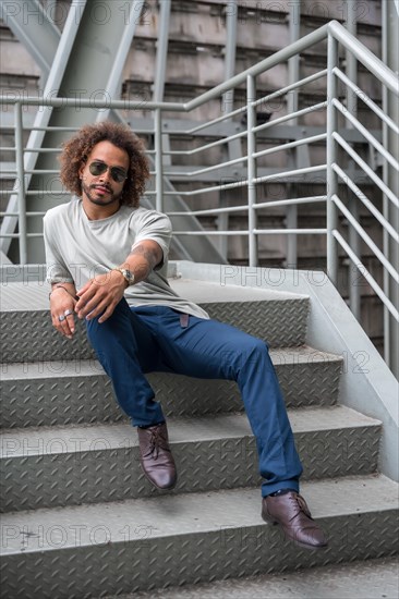 Portrait of a young man with afro hair wearing sunglasses on the stairs in the city sitting