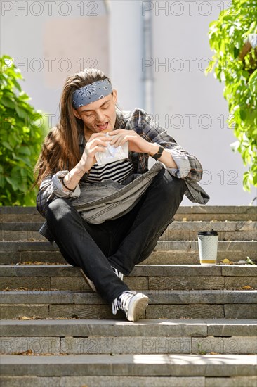 Long-haired man sits on staircase and eats croissant
